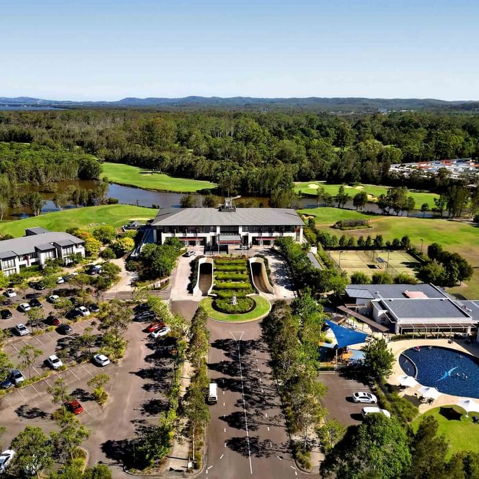 Aerial view of a pool with people swimming, surrounded by loungers, grass, and a parking lot at Mercure Kooindah Waters