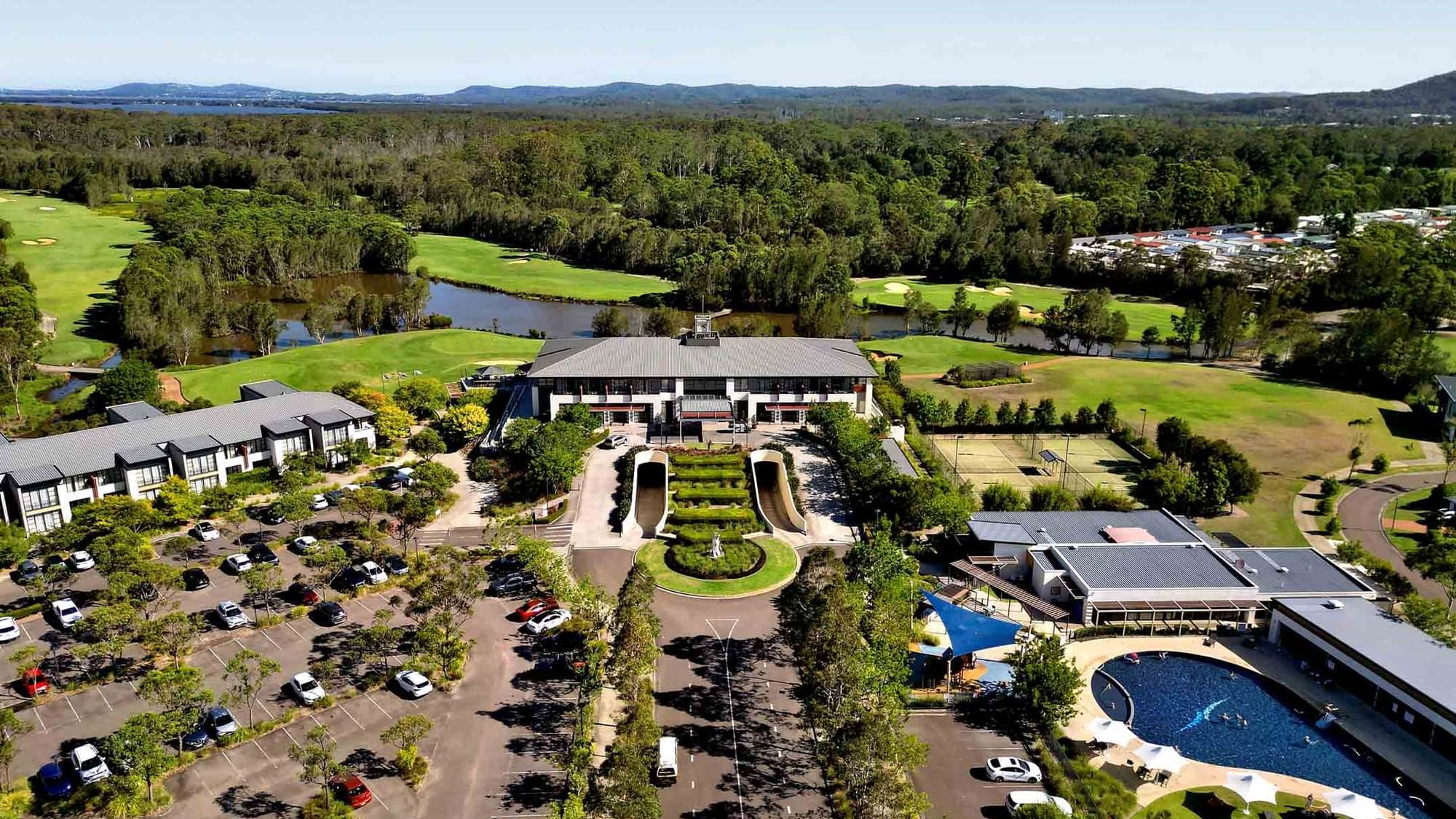 Aerial view of a pool with people swimming, surrounded by loungers, grass, and a parking lot at Mercure Kooindah Waters