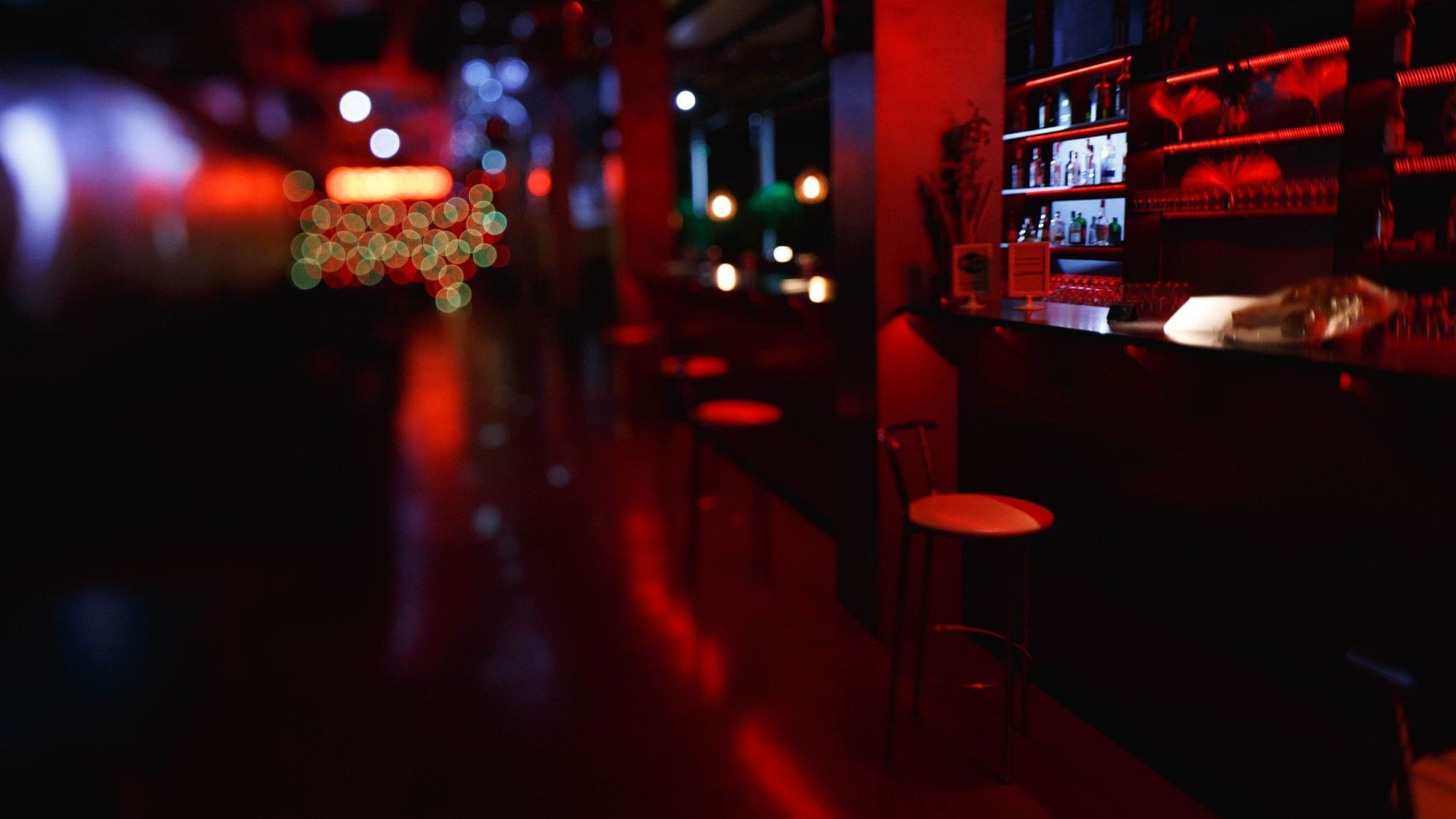 A dimly lit bar with red lighting, shelves of liquor bottles, and several bar stools.