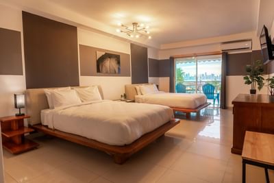 Two beds facing TV and balcony seating area in Deluxe Double at MARINN Tropical Vibes Hotel