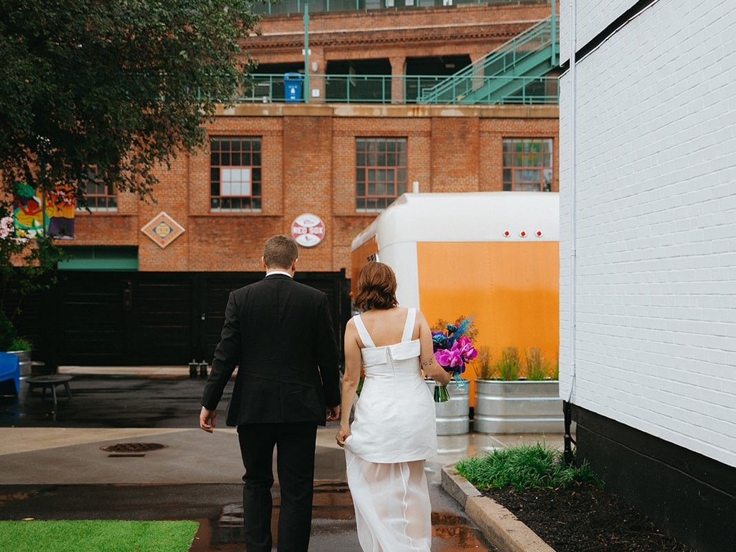 Couple in wedding attire walking hand in hand toward a colorful urban venue with vibrant bouquet and modern signage.