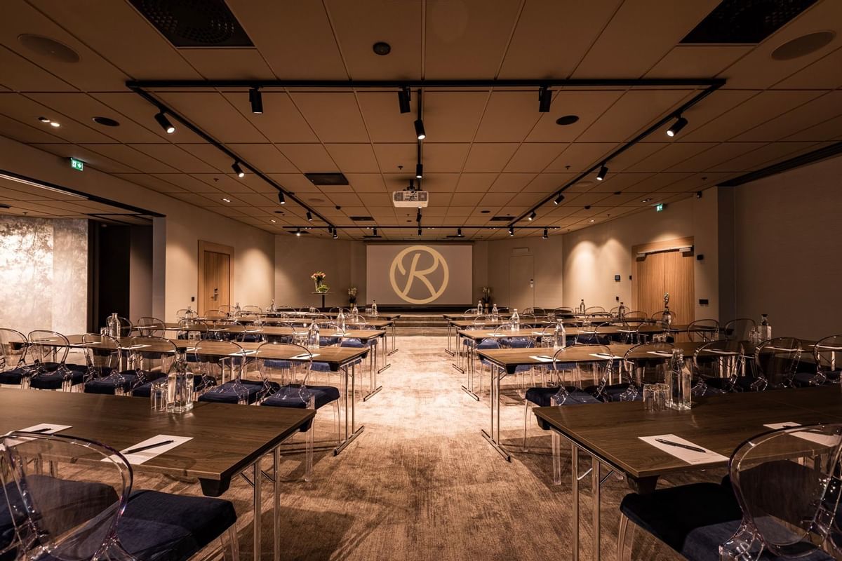 Konferensrum med bord och stolar, projektor och en stor bokstav R på väggskärmen.