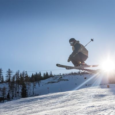 Skifahrer in der Luft über einem verschneiten Hang mit Sonnenstrahlen und Hintergrund von Bäumen und Liften.