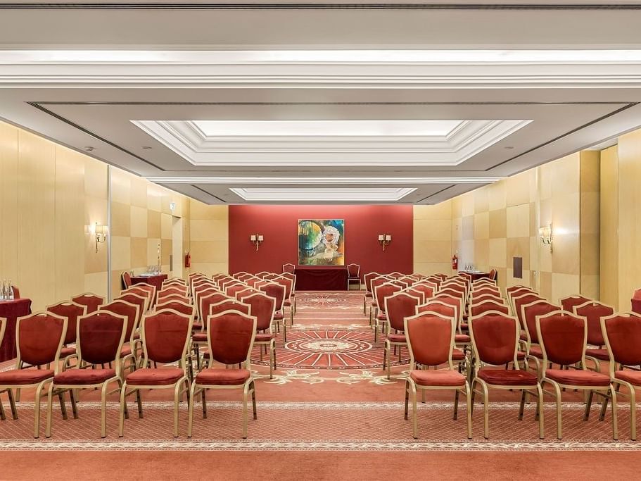 Sala de conferencias con sillas rojas, mesas cubiertas de rojo y pintura abstracta en la pared.