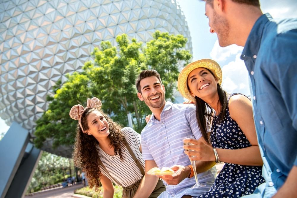 Four adults smile at one another holding food and a glass of Champagne in front of the iconic Spaceship Earth globe at EPCOT. 