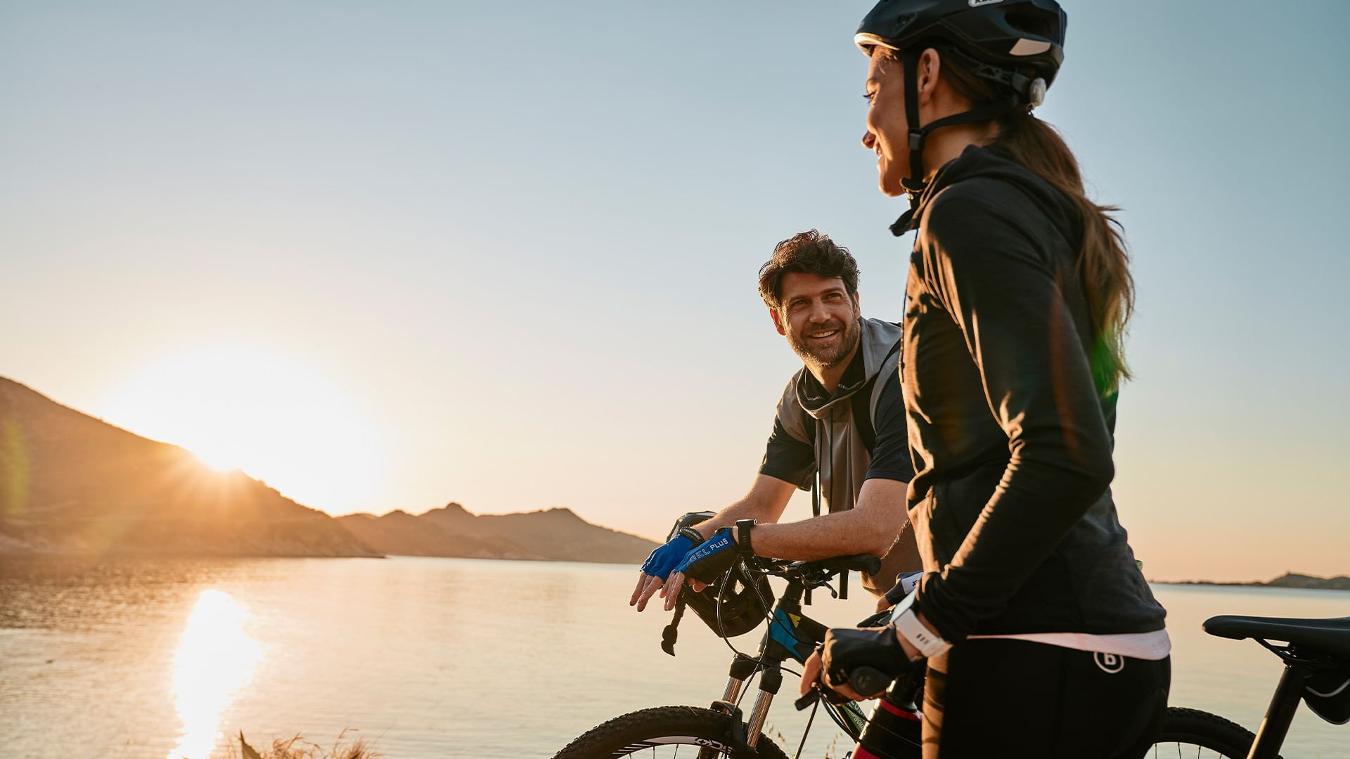 Zwei Radfahrer mit Helmen und Handschuhen unterhalten sich am Ufer eines Sees bei Sonnenuntergang.