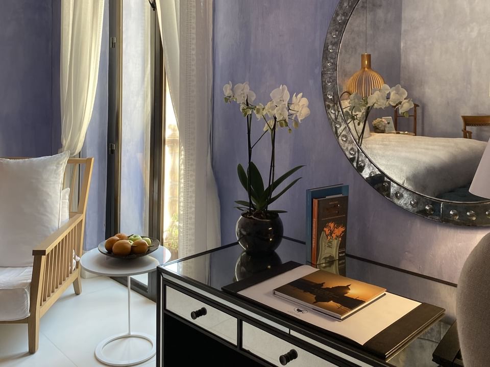Charleston Santa Teresa room with a glass desk, a vase with flowers, a round mirror, and a chair with a pillow.