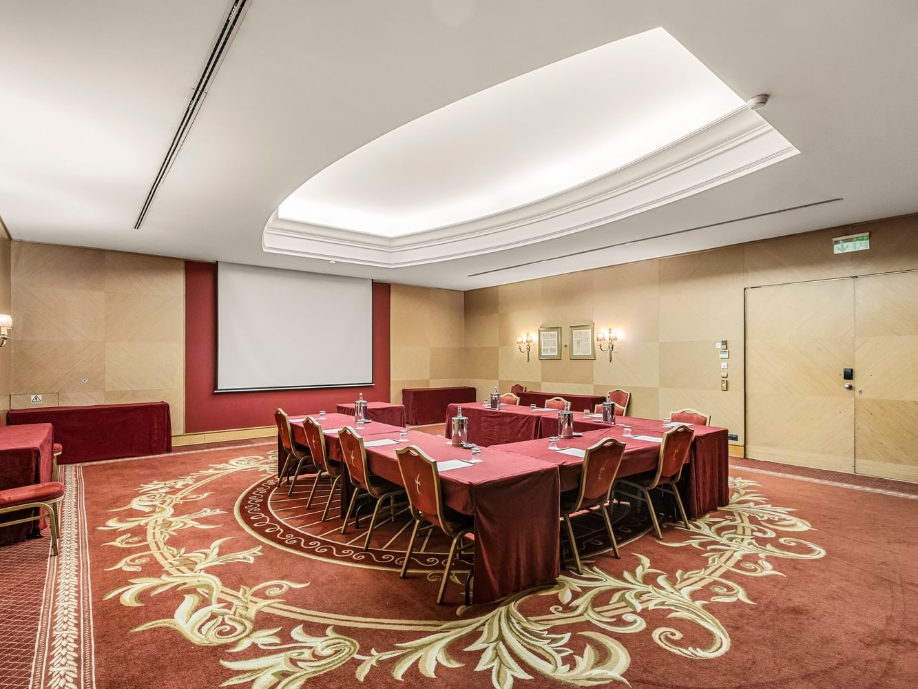 Sala de conferencias con mesas y sillas cubiertas en rojo, pantalla y paredes decoradas.