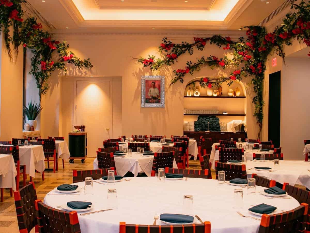 A dining room with round tables set for a meal, decorated with red floral arrangements and a high ceiling.