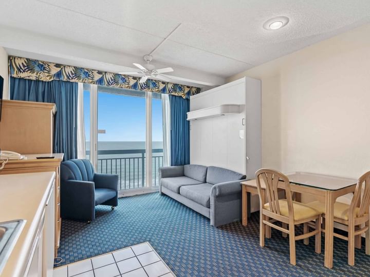 A wide view of a hotel room with a sofa, dining table, balcony, and kitchenette.