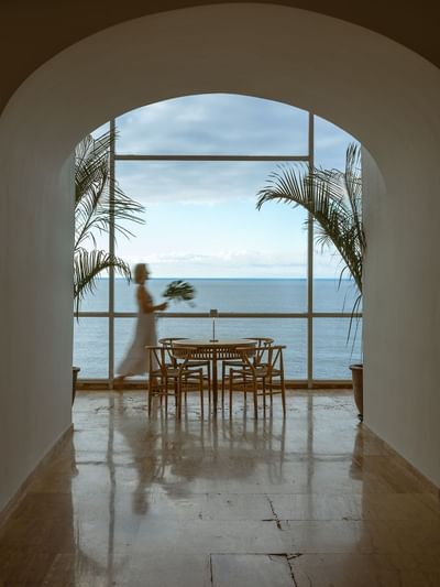 A woman walks past a dining table towards an ocean view through large windows.
