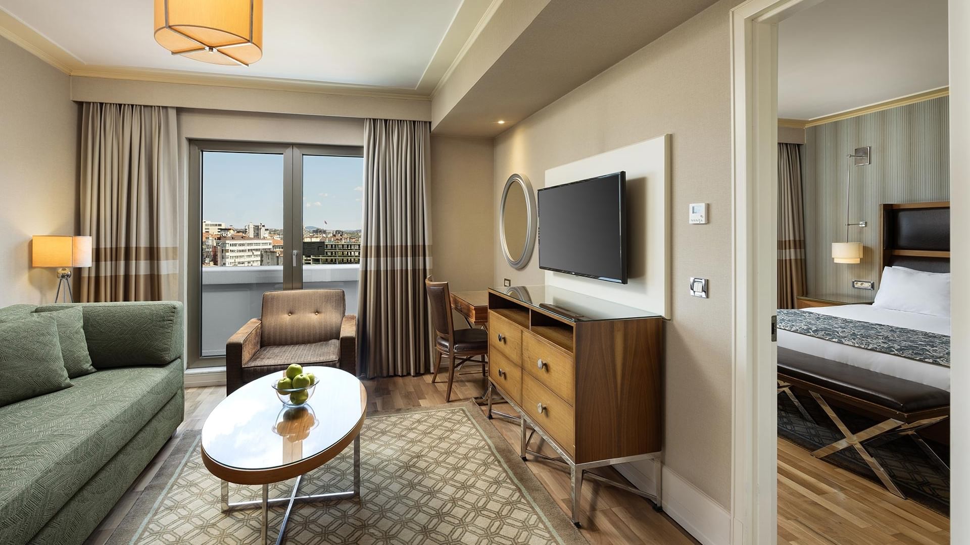 Living room with green couch, glass table, wooden dresser, TV, and view of a city through a large window in a suite at Titanic City Taksim.