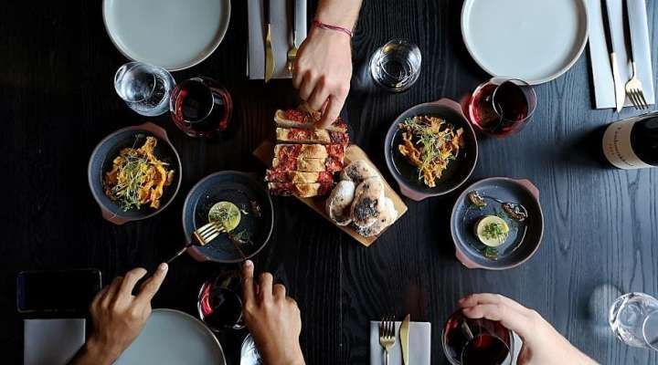 Several hands reaching for food on a dark table with plates, cutlery, wine glasses, and a bottle at Pepperclub Hotel