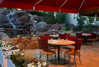 An outdoor patio dining area in Cascades Restaurant with red umbrellas and a rock waterfall at The Stanley Hotel