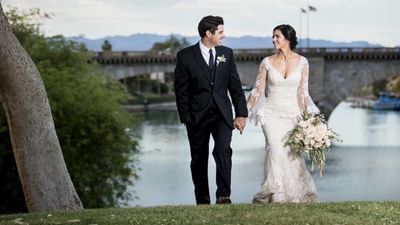 Bride and groom holding hands on waterfront lawn with bridge and mountains in background at London Bridge Resort