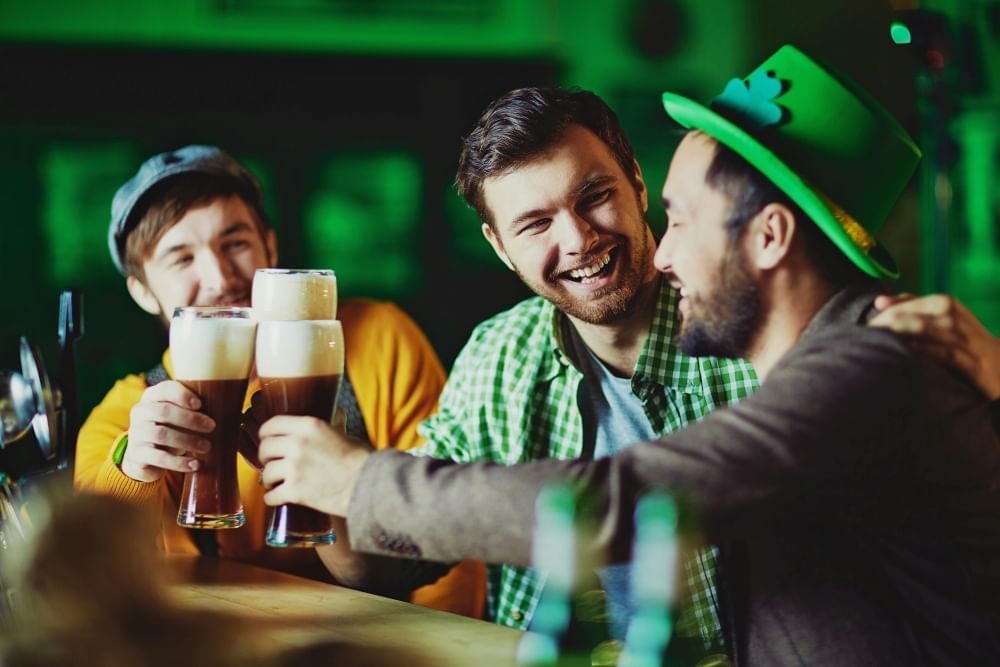 Three men, one in a green hat with a shamrock, toast three tall pints of dark beer while smiling.