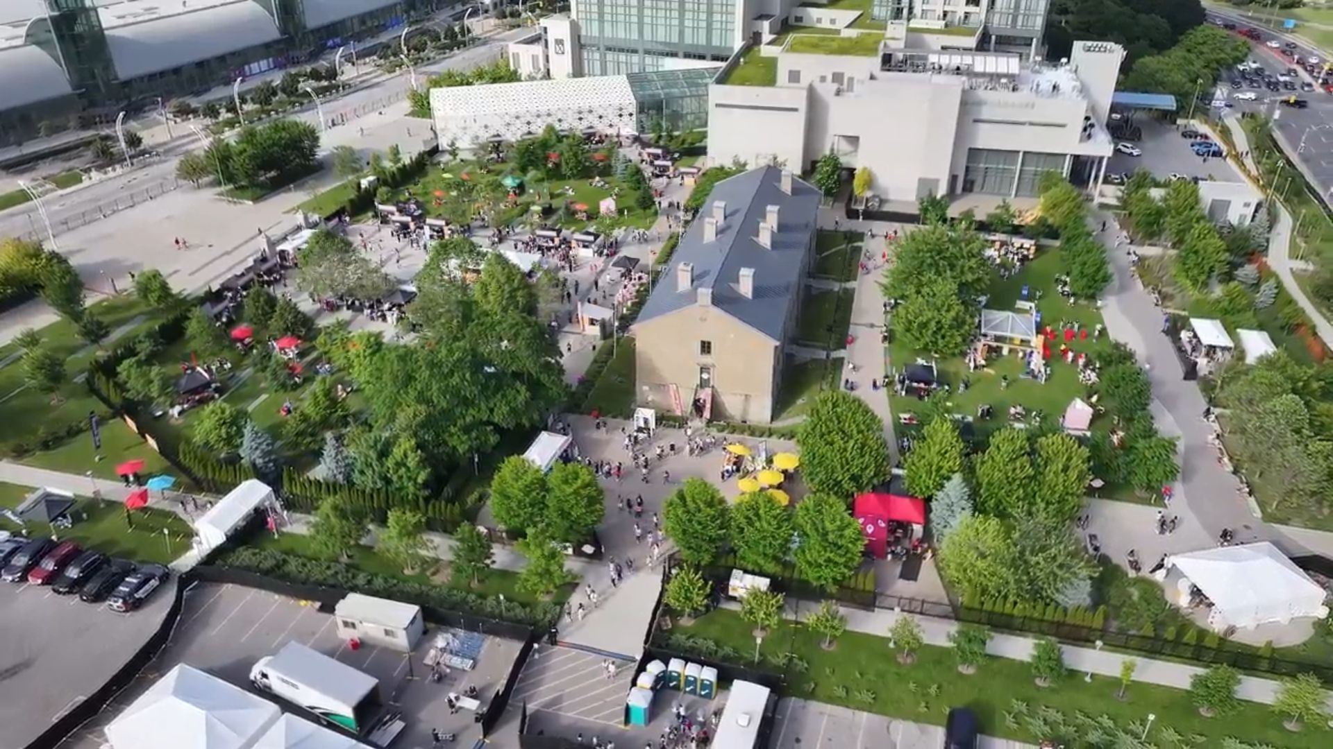 Aerial view of a festival with striped tents and crowds on green lawns, surrounded by trees near Hotel X Toronto
