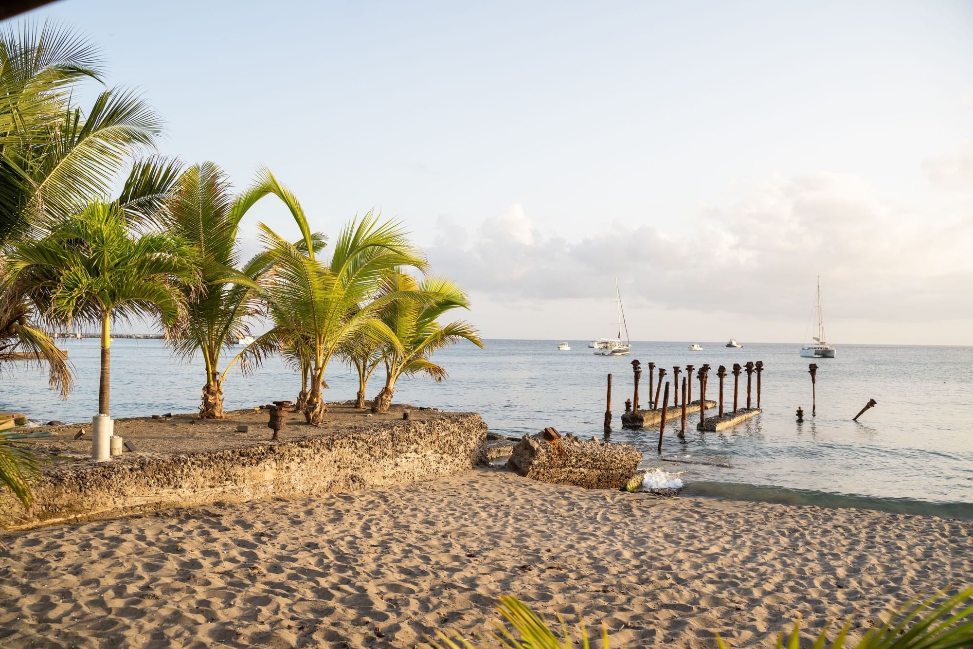 Sailboats floating on calm blue ocean water past old, weathered wooden pier ruins at Golden Rock Resort