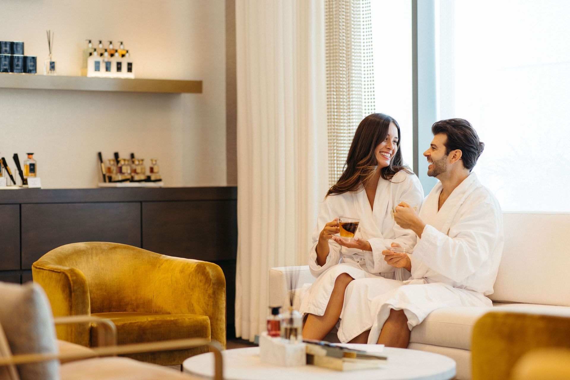 Couple in white bathrobes enjoying tea on a sofa between velvet mustard yellow armchairs at Guerlain Spa Hotel X Toronto