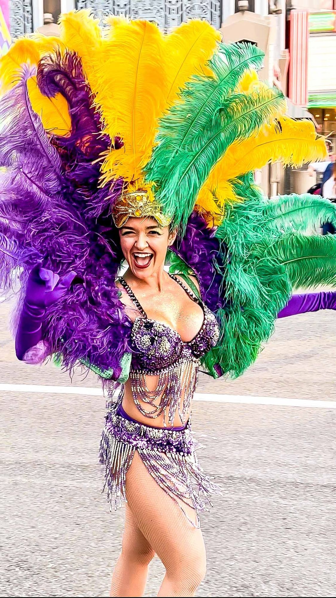 A performer in a purple crystal bikini with purple gloves and an enormous feathered headdress in purple, green, and yellow. 