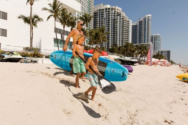 Father and two boys carry kayaks along a sunny, sandy beach near The Diplomat Resort