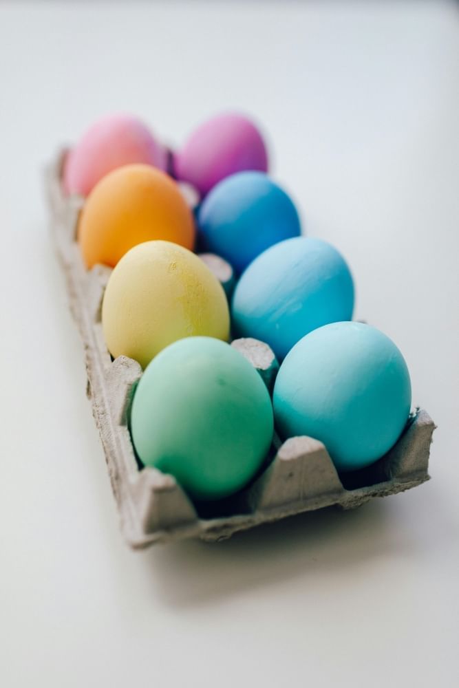 A cardboard egg carton with eight colorfully dyed eggs on a white surface.