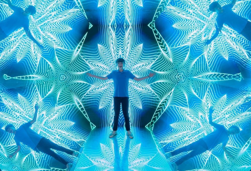 A boy stands in the middle of a blue glowing kaleidoscope exhibit. 