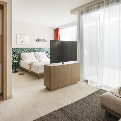 Camera d'albergo moderna con letto, TV, divano, tavolino e finestra con tende bianche.