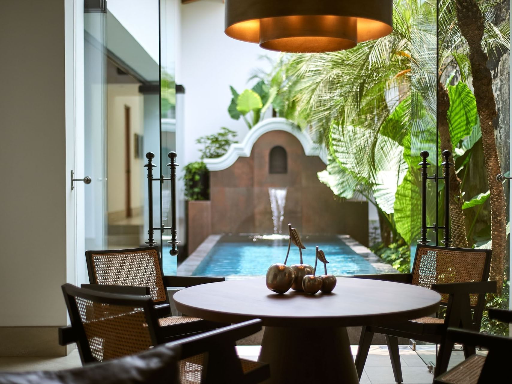 Round dining table with four chairs and decorative fruit centerpiece, overlooking a pool and lush garden.
