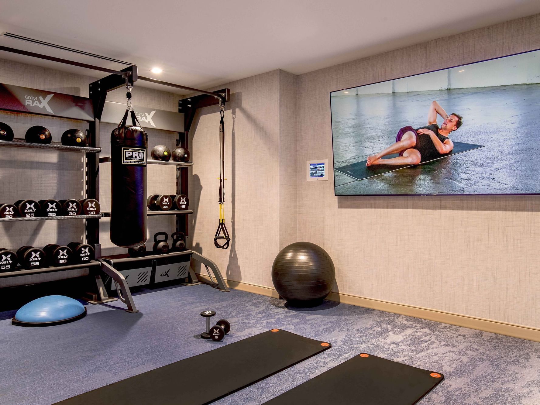 A fitness room with workout equipment, a large screen showing a person exercising, and exercise mats on the floor.