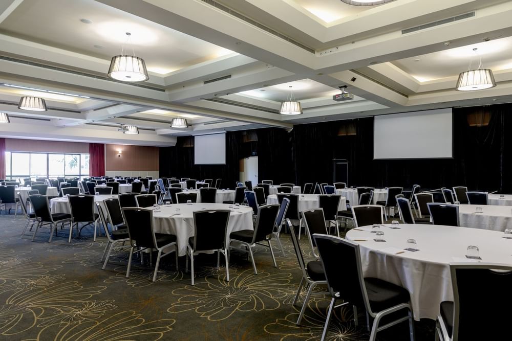 Banquet hall with round tables and black chairs, white tablecloths, ceiling lights, and two large screens on the wall.