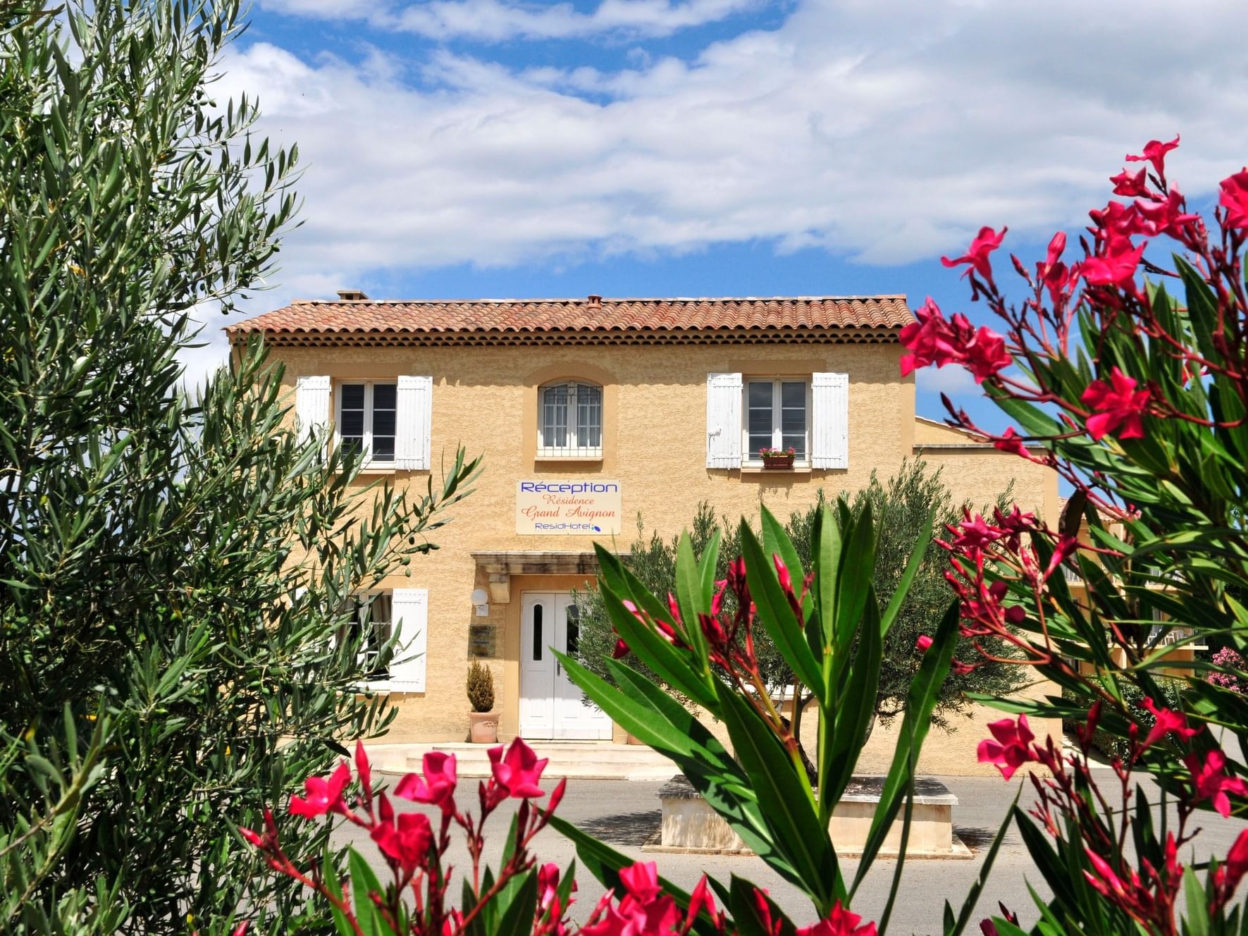 Exterior of ResidHotel Grand Avignon framed by vibrant pink flowers and lush greenery under a partly cloudy sky
