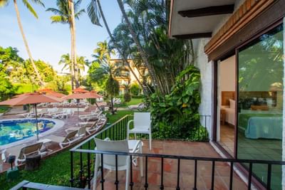 Patio view of resort with pool, lounge chairs, umbrellas, and lush greenery under clear sky.