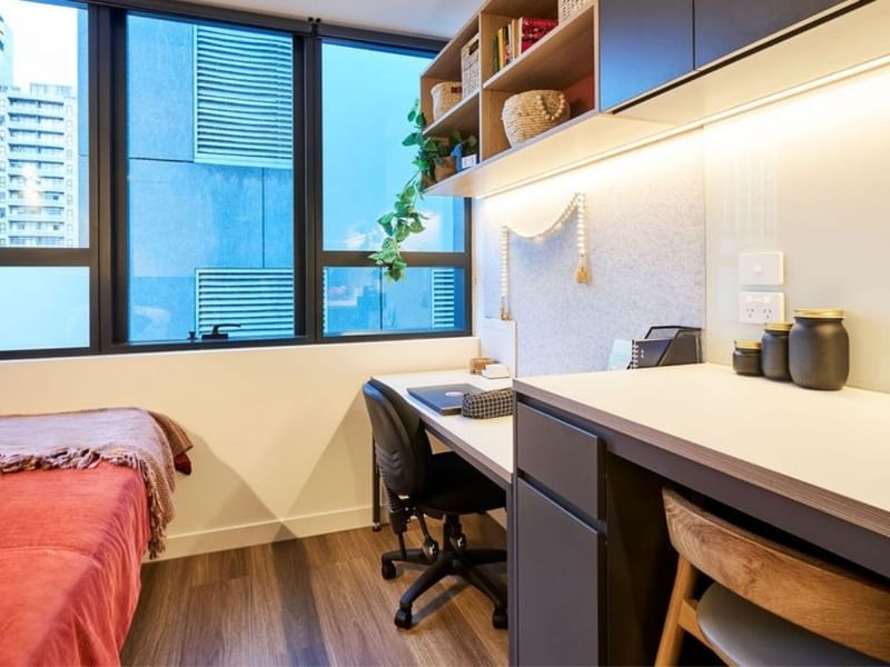 Modern bedroom with desk, chair, built-in shelves, and large window overlooking cityscape.