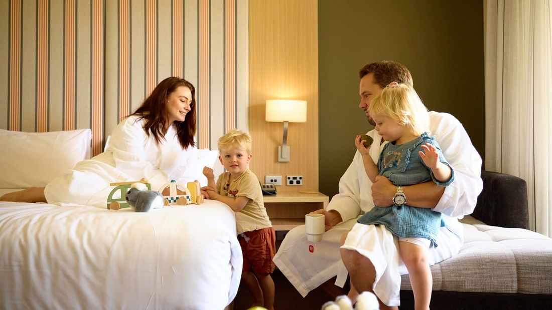 Family of four in bathrobes in a hotel room, with children playing with toys and fruit on a table.