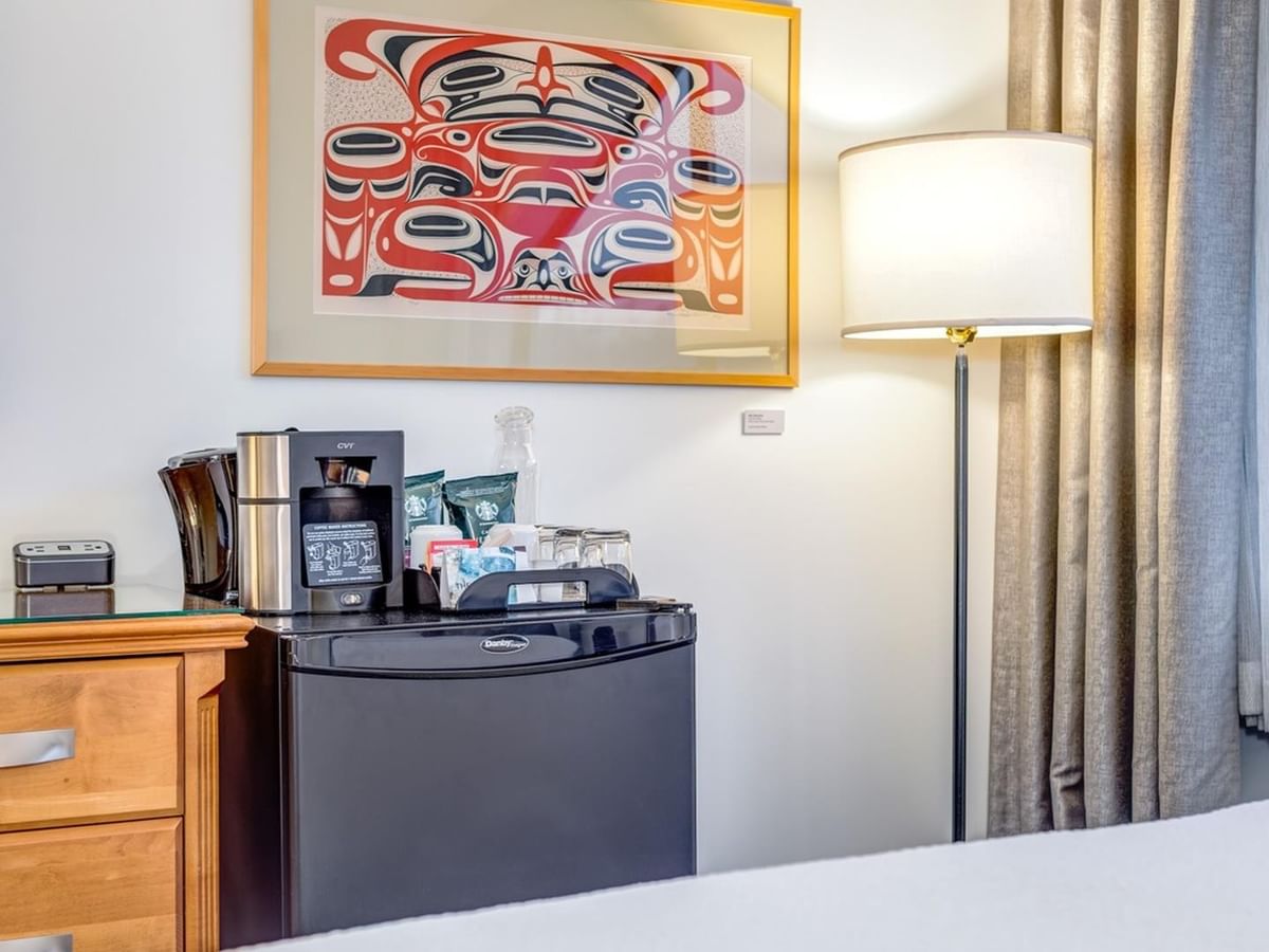 Room with a bed, a mini-fridge with coffee maker, a lamp, and a framed artwork.