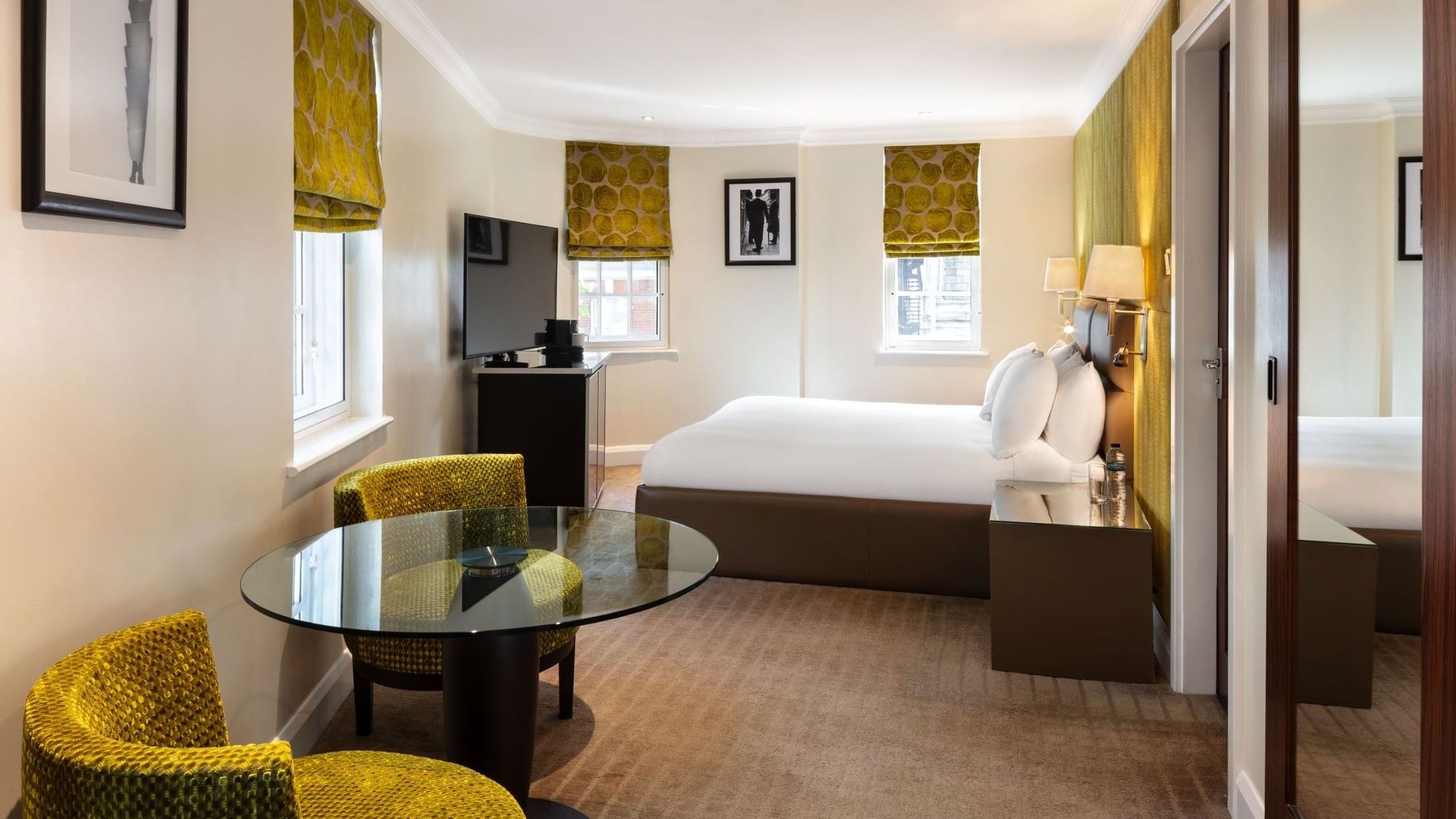 Deluxe Room at The May Fair Hotel with a large bed, yellow-patterned blinds, green chairs, a glass table, and warm lighting