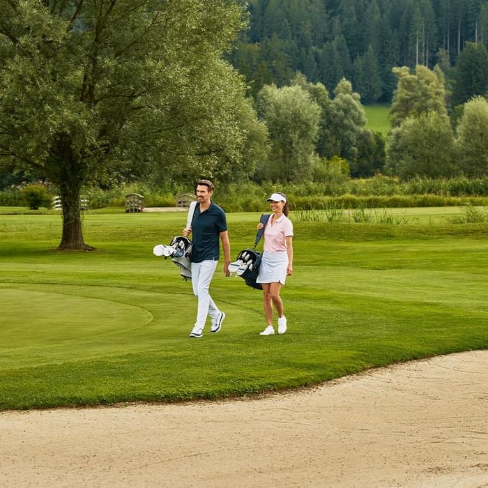 Paar mit Golfschlägern auf einem Golfplatz, umgeben von Bäumen und Sandbunker im Vordergrund.