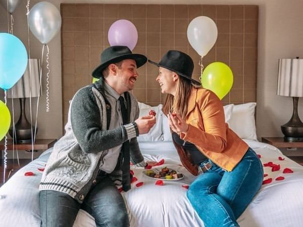 Couple on bed with balloons, woman holds food, man looks at her, both wear hats.