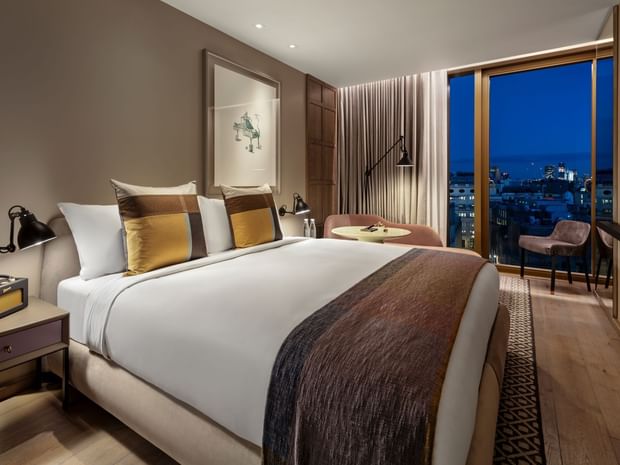 Bed by the large windows with city views in Deluxe King City View Room at The Londoner Hotel