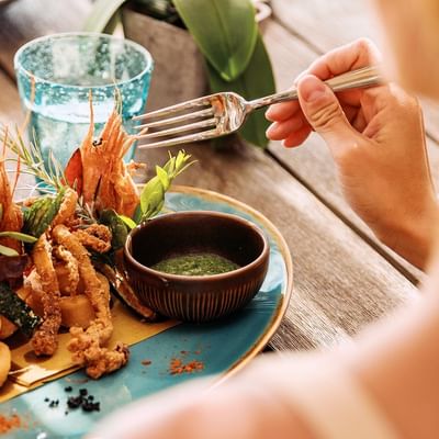 Person mit Gabel vor einem Teller mit Garnelen, Tintenfischringen und Dip auf einem Tisch.