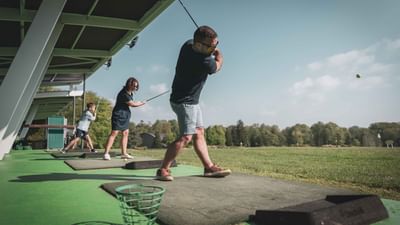 Three people practicing golf on a driving range with trees and a blue sky in the background.