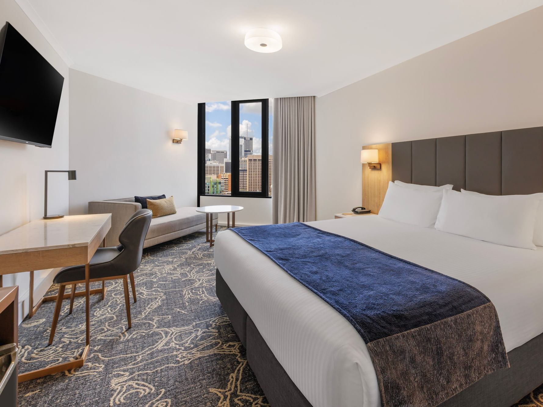 Modern king hotel room with city view, featuring a white bed with blue throw, executive work desk, and lounge seating.