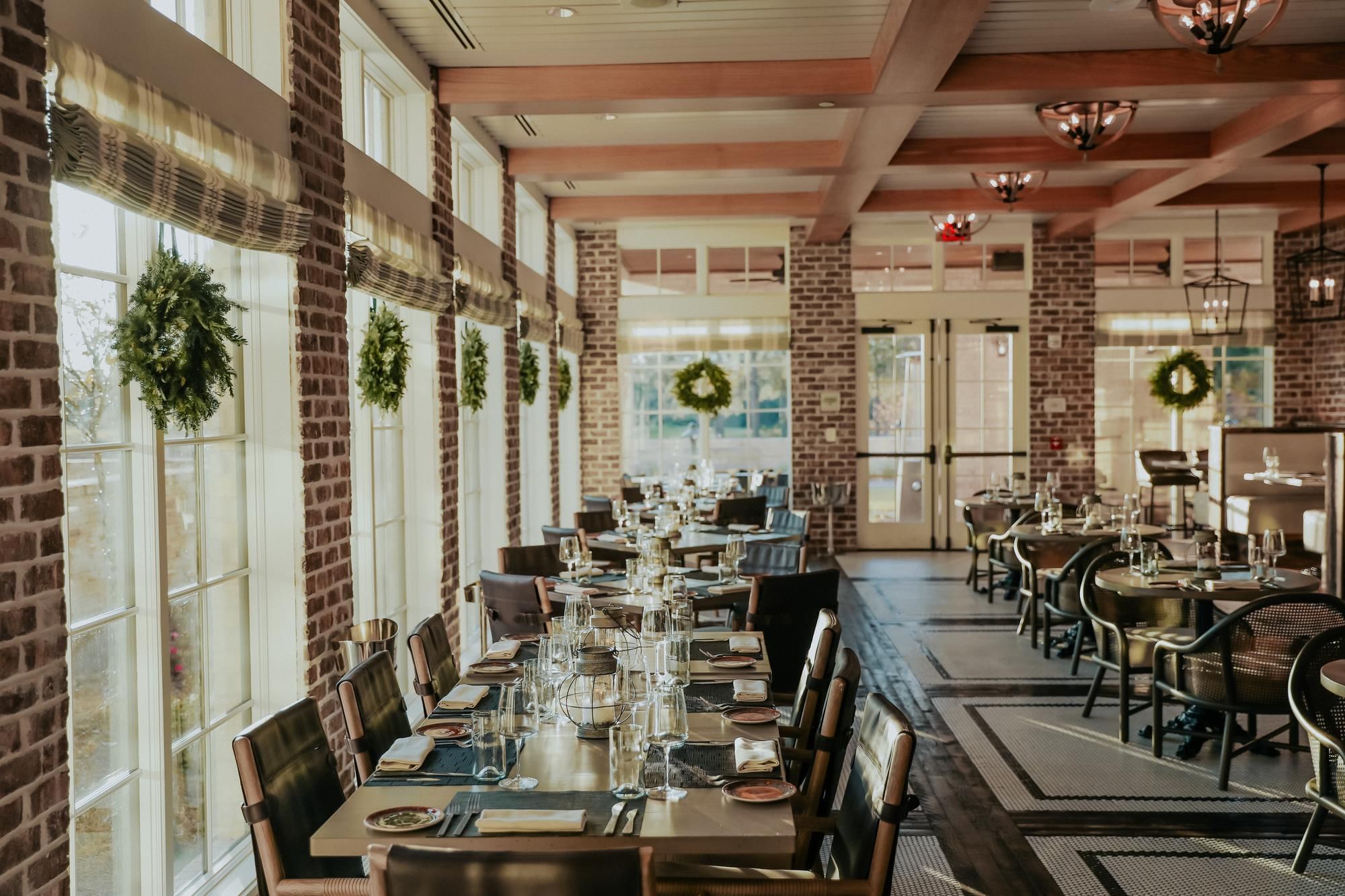 A brick-walled dining room with large windows, set tables, greenery decor, and wooden ceiling beams.