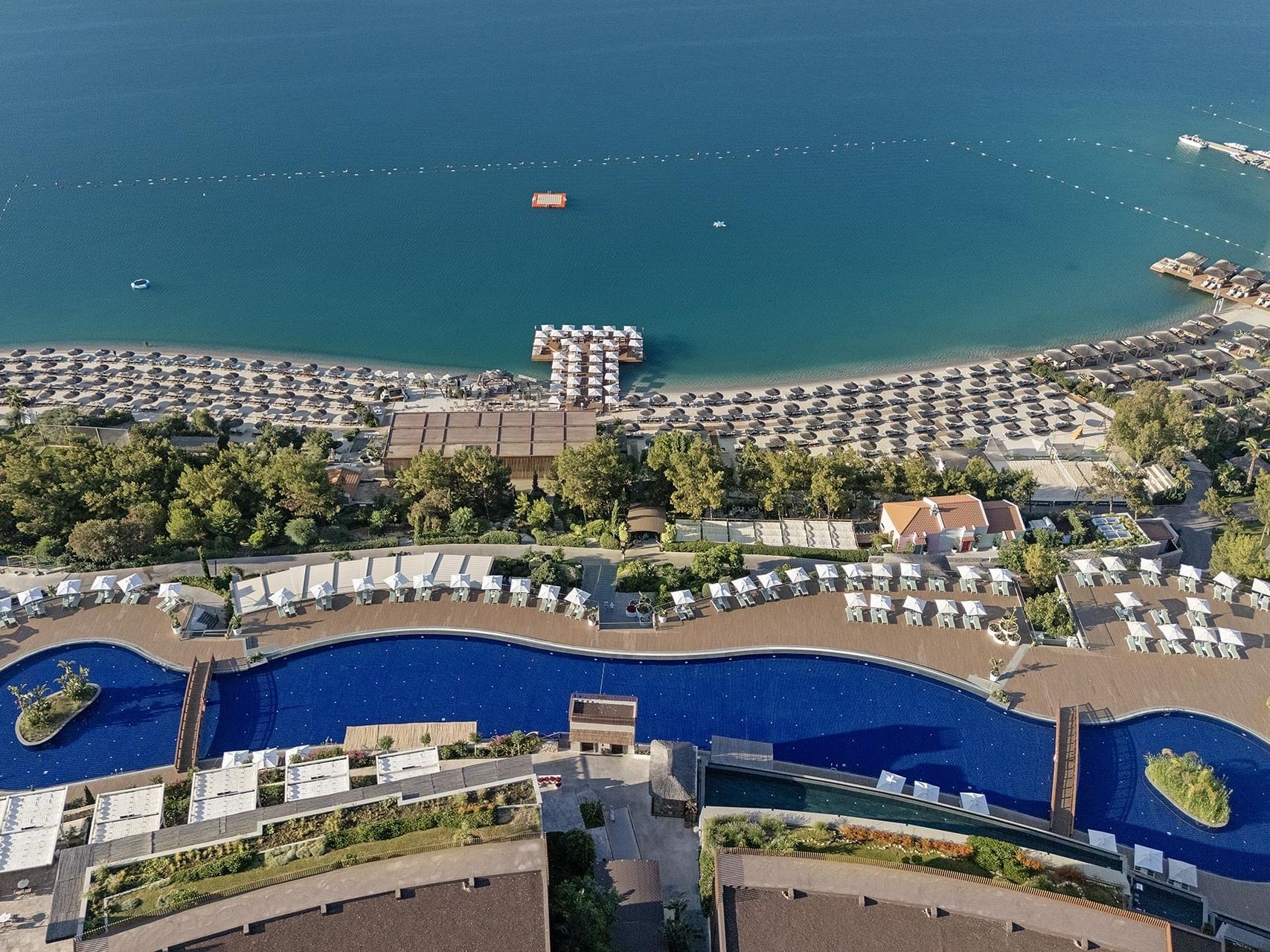 Aerial view of resort with blue pool, beach, and ocean, surrounded by trees and lounge chairs Titanic Luxury Collection Bodrum.