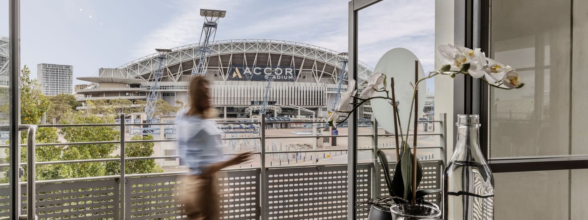 Blurred figure walking past a balcony table overlooking Accor Stadium in Marine Room at Novotel Sydney Olympic Park
