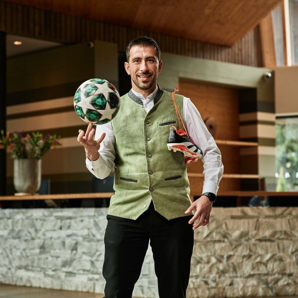 Mann in formeller Kleidung hält einen Fußball und trägt einen Fußballschuh an einer Schnur um den Hals.
