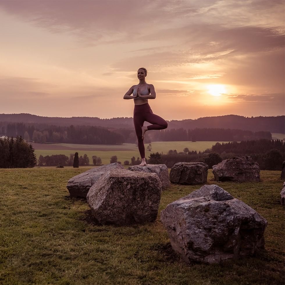 Donna che pratica yoga all'alba su rocce in un campo verde.