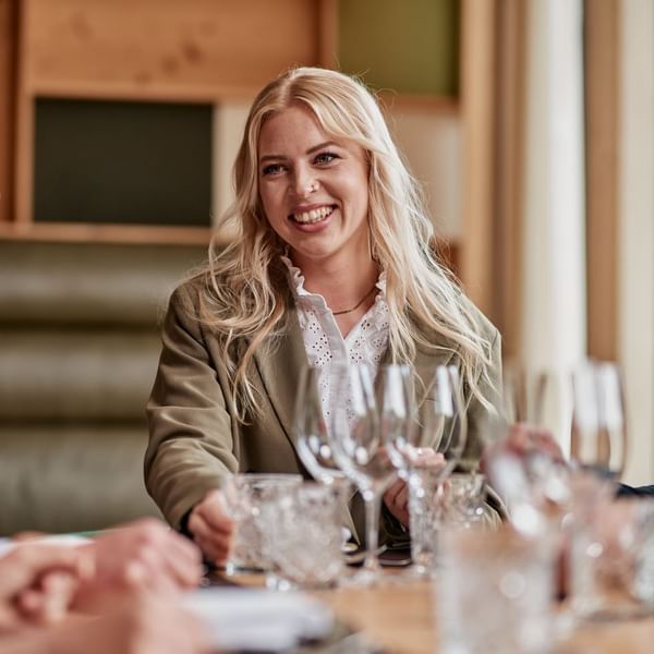 Eine Frau mit blonden Haaren sitzt an einem Tisch mit mehreren Gläsern und lächelt.