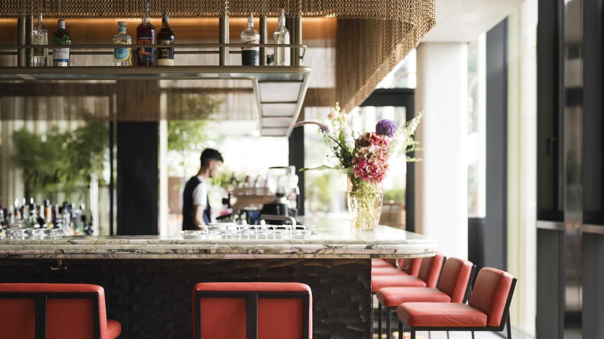 Bar con fiori in vaso e uomo al lavoro, sedie rosse, bottiglie di liquore e alcolici.